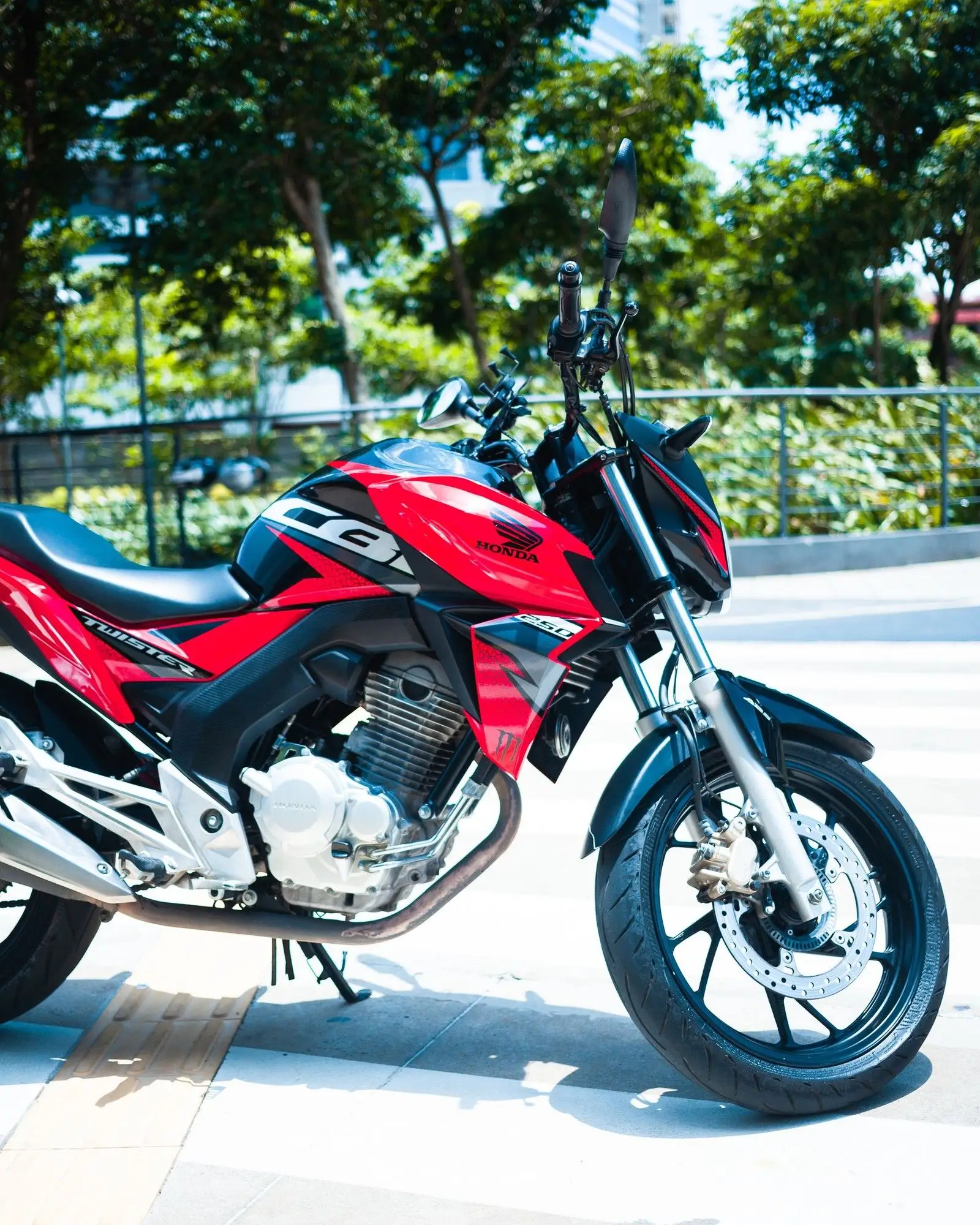 Moto de rua vermelha estacionada numa calçada, com árvores e prédios ao fundo